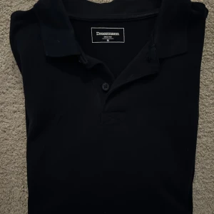 Svart pikétröja från Dressmann - Snygg svart pikétröja från Dressmann i storlek M. Tröjan har en klassisk krage och knappar framtill. Perfekt för en stilren look.