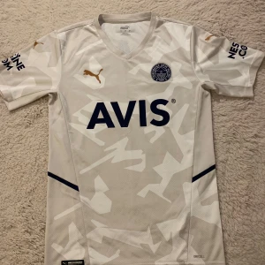 Fenerbahce borta 2022/23 - Snygg vit fotbollströja från Puma med kamouflagemönster och korta ärmar. Tröjan har guldiga Puma-loggor och sponsortryck i mörkblått. Köpt från Fenerbahce club store i Istanbul.