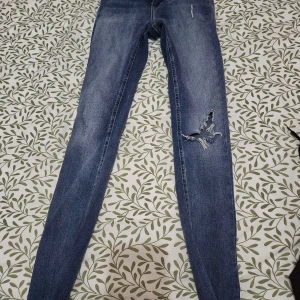 Blå jeans från ONLY - Snygga blå jeans från ONLY med slitna detaljer på ena benet. De har en klassisk femficksdesign och stängs med dragkedja och knapp. Perfekta för en avslappnad stil.