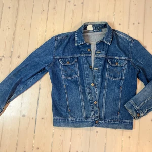 Blå jeansjacka från Wrangler - Vintage Wrangler Jeansjacka – Slitstark och Cool!  Säljer en klassisk Wrangler original jeansjacka i vintage-stil.   Slitstark och cool, perfekt för att få den autentiska 70-80-tals looken.    •   Design: Tidlös och robust med alla de rätta detaljerna för en äkta vintagelook.    •   Stil: Perfekt för våren, eller när du är ute och kör moped eller bara vill ha ett retroinspirerat uttryck.    •   Komfort: Bekväm passform för både vardagsbruk och lite tuffare aktiviteter.  En jacka som håller både 