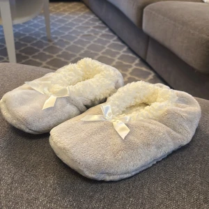 Gråa tofflor med rosett - Mysiga gråa tofflor med vit fluffig insida och en söt rosett på ovansidan. Perfekta för att hålla fötterna varma hemma. Sulan har små gummidetaljer för bättre grepp.(helt nya) 