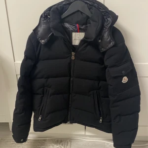 Moncler jacka  - Säljer en snygg svart dunjacka med dragkedja och avtagbar huva. Jackan har långa ärmar och praktiska fickor med dragkedjor. Perfekt för kyliga dagar.