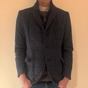 Mörkblå kavaj från Boggi Milano - Snygg mörkblå kavaj från Boggi Milano med quiltad design och knappar framtill. Perfekt för en stilren look. Kavajen har långa ärmar och en klassisk krage. Funkar bäst på hösten och våren men funkar även i en varmare vinter. En knapp saknas men går att sy på och hänger med kavajen på köp. Förutom det är kavajen i topp skick. Modellen är drygt 190 cm.