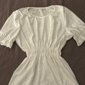 Vit blouse med spetsdetaljer - Charmig vit blouse med spetsdetaljer och elastisk midja. Den har korta ärmar och en rund halsringning, perfekt för sommarens varma dagar. Blousen är både söt och bekväm, vilket gör den till ett måste i garderoben.