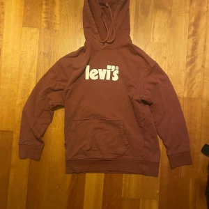 Vinröd hoodie från Levi's - Säljer en snygg vinröd hoodie från Levi's med vit logga på bröstet. Den har en klassisk känguruficka och justerbar huva. Perfekt för en avslappnad stil.