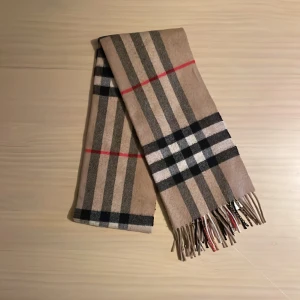 Burberry halsduk  - Säljer en halsduk från Burberry i beige, svart och röd. 100% kashmir. Den är äkta men har inte kvar kvittot då jag fått den som en gåva.