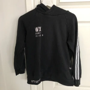 Svart hoodie med tryck och ränder - Säljer en svart hoodie från WX med tryck på bröstet och ryggen. Tröjan har långa ärmar med vita ränder längs sidorna och en bekväm huva. Perfekt för en avslappnad stil.