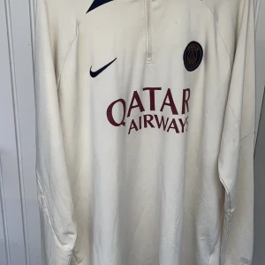 Beige långärmad fotbollströja från Nike ( Paris saint german tröja) - Snygg beige långärmad fotbollströja från Nike med Paris Saint-Germain-logga och Qatar Airways-tryck på framsidan. Perfekt för fans av PSG! Tröjan har en dragkedja vid halsen för extra komfort.