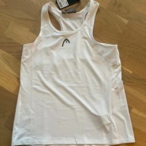 Vit sport linnetopp från Head -Dam - Säljer en vit linnetopp från Head i syntetmaterial. Perfekt för träning med sin bekväma passform och stilrena design. Toppen har ett diskret logotryck på framsidan och brottarrygg för extra rörelsefrihet. Den är för dam och helt ny, nypris ca 400kr
