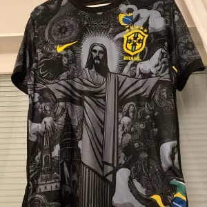 Svart Brasilien fotbollströja med Kristusmotiv - Snygg svart fotbollströja från Nike med Brasilien-emblem och ett stort Kristusmotiv på framsidan. Tröjan har korta ärmar med detaljer i gult och blått. Perfekt för fotbollsfans som vill sticka ut.