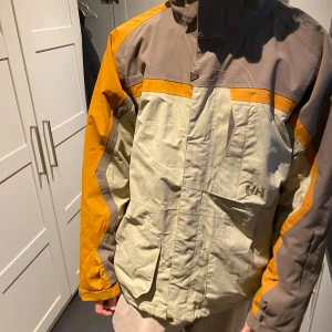 Flerfärgad jacka från Helly Hansen - Säljer en flerfärgad jacka från Helly Hansen med dragkedja och hög krage. Jackan har en kombination av beige, brun och orange färgblockdesign. Perfekt för utomhusaktiviteter med praktiska fickor och justerbara ärmslut.