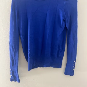 Blå långärmad tröja med knappar - Snygg blå långärmad tröja med dekorativa knappar vid ärmsluten. Perfekt för en stilren look.