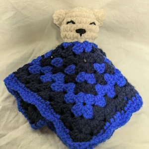 Virkad snuttefilt med björnmotiv - En söt virkad snuttefilt i blå och svart med ett björnansikte i beige. Perfekt för små barn att mysa med. Filten har ett mönster av stora rutor och är handgjord med kärlek.