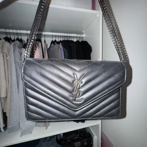 Silver axelväska från YSL - Snygg silverfärgad axelväska (inte äkta YSL )mycket bra skick. Väskan är quiltad med ett elegant mönster och har en kedjerem. Perfekt för både i vardag och fest! Insidan har en praktisk dragkedja för säker förvaring.