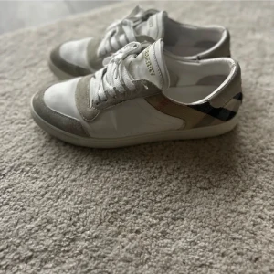 Vita sneakers från Burberry - Snygga vita sneakers från Burberry med klassiskt rutigt mönster på hälen. Skorna har snörning och detaljer i mocka som ger en stilren look. Perfekta för dig som vill ha en riktigt clean look men vill även sticka ut från mängden! 