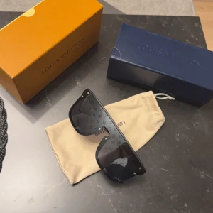 Svarta solglasögon från Louis Vuitton - Snygga svarta solglasögon från Louis Vuitton med ett diskret mönster på glasen. De har en modern design med breda skalmar och små nitar i hörnen. Perfekta för en stilren look.