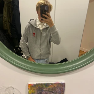 Grå hoodie med rött hjärta från Ami Paris - Säljer en stilren grå hoodie från Ami Paris med ett rött hjärta och bokstaven 'A' broderat på bröstet. Tröjan har dragkedja och en bekväm huva. Perfekt för en avslappnad look. Priset går att diskuteras! Nypris 3790 kr! Storlek mellan S och M men passar bra på båda.