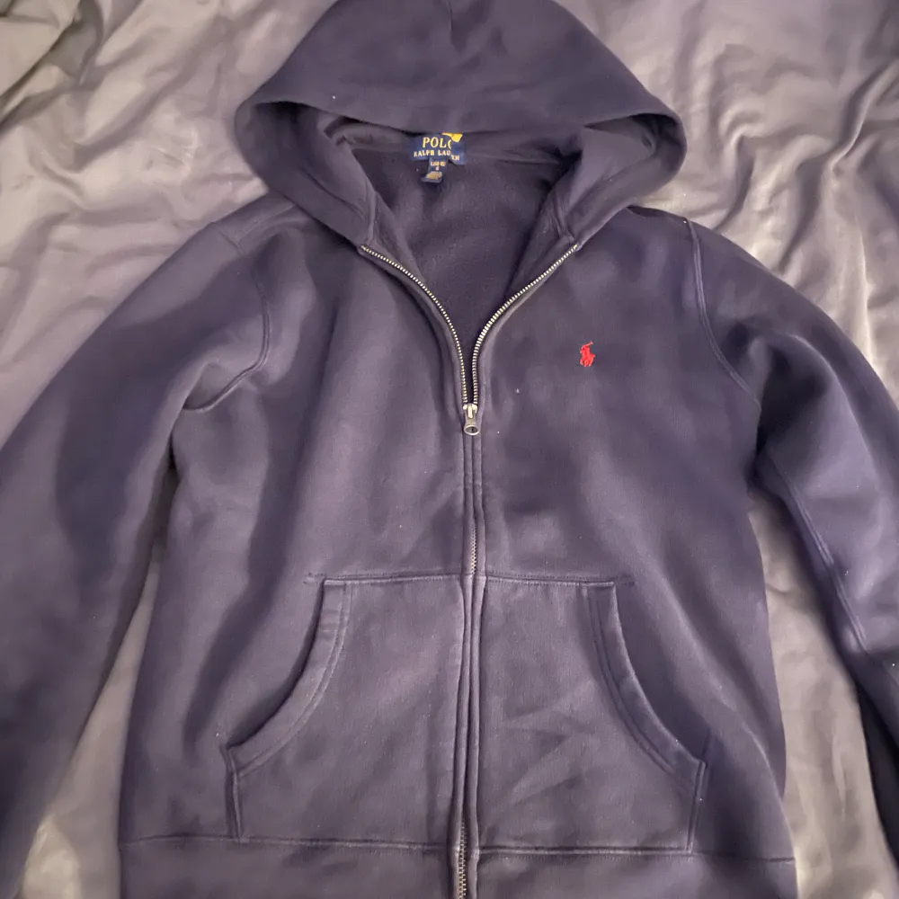 Säljer en snygg mörkblå hoodie från Ralph Lauren med dragkedja och röd logga på bröstet. Tillverkad i en mjuk bomullsblandning med fickor framtill. Perfekt för en avslappnad stil.. Hoodies.