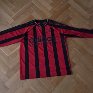 Röd och svart randig träningströja från Adidas - Snygg långärmad fotbollströja från Adidas i röd och svart randigt mönster. Perfekt för fotbollsälskare som vill ha en klassisk look på planen. Tröjan har en bekväm passform och är tillverkad i ett lätt material.