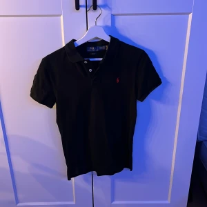 Svart kortärmad pikétröja från Ralph Lauren - Säljer en stilren svart pikétröja från Ralph Lauren med röd broderad logga på bröstet. Tröjan har en slim fit passform och korta ärmar, perfekt för en klassisk look.