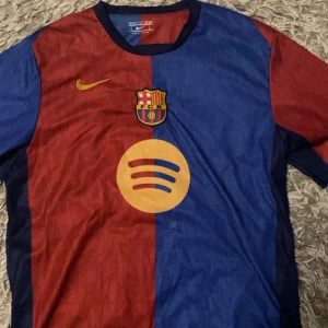 FC Barcelona fotbollströja - Säljer en FC Barcelona tröja i rött och blått från Nike. Den har korta ärmar och klubbens emblem på bröstet. På framsidan finns en stor Spotify-logga och på baksidan står det 'Lamine Yamal' med nummer 19. Perfekt för fans som vill visa sitt stöd!