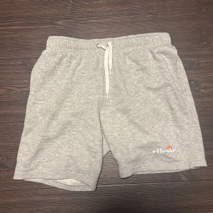 Grå shorts från Ellesse - Säljer ett par grå shorts från Ellesse i mycket bra skick. De är gjorda av mjuk bomull och har en bekväm passform med resår och snörning i midjan. Perfekta för avslappnade dagar eller träning. Loggan är broderad på ena benet.