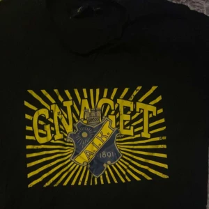Svart t-shirt med AIK-tryck - Säljer en svart t-shirt med ett stort gult och blått AIK-tryck på framsidan. Perfekt för alla AIK-fans som vill visa sitt stöd. T-shirten är i bra skick och passar utmärkt till vardags eller på matchdagar. Skön och bekväm att bära!