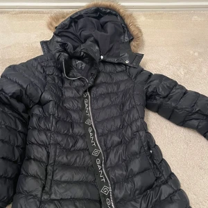 Svart dunjacka med pälskrage från Gant - Snygg svart dunjacka från Gant med pälskrage och dragkedja. Perfekt för kalla dagar med sin varma och stilrena design. Jackan har långa ärmar och en praktisk huva.