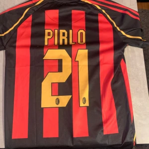AC Milan fotbollströja med Pirlo 21 storleken är S, färgerna röd svart guld, kom med frågor med mera - Säljer en klassisk AC Milan fotbollströja från Adidas i svart och rött med gula detaljer. Tröjan är kortärmad och har sponsorloggan 'bwin' på framsidan. På baksidan står det 'Pirlo 21'. Perfekt för samlare eller fans av laget! Storleken är S, färgerna röd svart guld, kom med frågor med mera