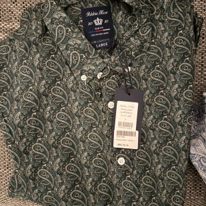 Helt ny Grön paisleymönstrad skjorta från Robbie Moor - Snygg grön skjorta från Robbie Moor med paisleymönster. Skjortan har en slim fit passform och knappar framtill. Perfekt för en stilren look.