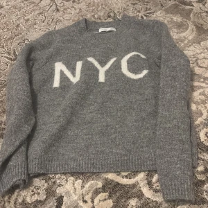 Grå tröja med NYC-text från Primark - Säljer en mysig grå tröja från Primark med vit text 'NYC' på framsidan. Tröjan har långa ärmar och är perfekt för kyligare dagar. Enkel och stilren design som passar till det mesta.