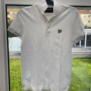 Vit kortärmad pikétröja från Lyle & Scott - Säljer en stilren vit pikétröja från Lyle & Scott i mycket bra skick. Den har en klassisk krage med knappar och en broderad logga på bröstet. Perfekt för både vardag och fest! 🕊️
