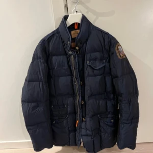 Mörkblå pufferjacka från Parajumpers - Säljer en snygg mörkblå pufferjacka från Parajumpers i bra skick utöver att det finns två hål. Ett fram och ett litet bak som knappt syns när man bär jackan. Jackan har en quiltad design med dragkedja och knappar framtill. Den har långa ärmar och en cool patch på ärmen. Perfekt för höst och vinter!