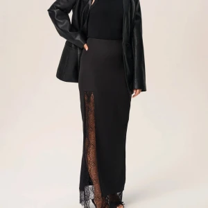 Svart långkjol med spetsdetalj från ADOORE - Säljer kjolen Spezia från ADOORE. Kjolen har en stilren design med en slits som är dekorerad med vacker spets. Den är i nyskick, storlek 34. 