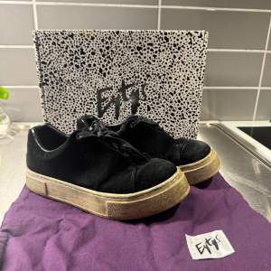 Snygga svarta sneakers från Eytys med en robust sula och klassisk snörning. Skorna har en stilren design med en liten lila detalj på hälen. Perfekta för en trendig look.