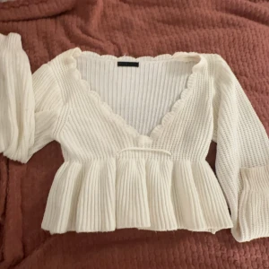 Vit ribbad omlottopp - Säljer en söt vit ribbad omlottopp med volangdetaljer. Toppen har långa ärmar och en v-ringad design som ger en feminin touch. Perfekt för en stilren look.