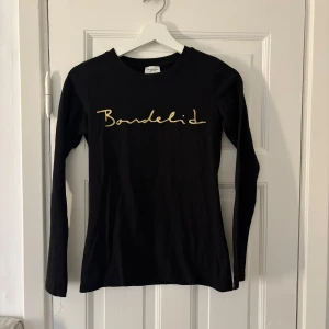 Svart långärmad tröja från Bondelid - Snygg svart långärmad tröja från Bondelid med guldfärgad logotyp på bröstet. Perfekt för en stilren look. Tröjan är i en bekväm passform och passar till många olika outfits.