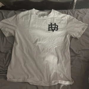 Vit t-shirt från Bvaldi - Säljer en vit t-shirt från Bvaldi med ett svart broderat märke på bröstet. T-shirten är kortärmad och har en klassisk rund halsringning. Perfekt för en avslappnad stil. Använd 7 gånger max