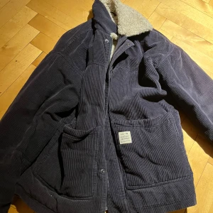 Blå manchesterjacka från Urban Outfitters - Säljer en snygg blå varm vinter manchesterjacka från Urban Outfitters med mysigt teddyfoder. Jackan har en klassisk krage och praktiska fickor framtill. Perfekt för kyliga dagar!