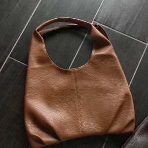 Brun axelväska i syntet - Säljer en snygg brun axelväska i syntetmaterial. Väskan har en enkel och stilren design med en bred axelrem som gör den bekväm att bära. Perfekt för vardagsbruk och rymmer det mesta du behöver ha med dig. Passar till alla säsonger och outfits!