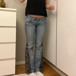 Jeans med leoparddetaljer - Snygga ljusblå jeans med unika leopardmönstrade detaljer längs sidorna. Byxorna har en bootcut-stil och är lågmidjade, vilket ger en trendig look. Perfekta för dig som vill sticka ut med en djärv stil. Mått: midja rakt över- 38 cm innerbenslängd- 73,5cm