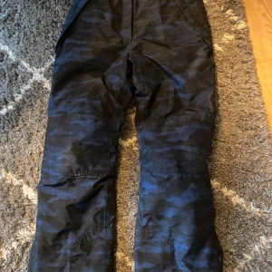 Camouflagefärgade täckbyxor från Everest - Säljer ett par svarta och blåa täckbyxor från Everest. De är vattentäta med TCS Waterproof-teknologi och har en vattenpelare på 3000 mm. Perfekta för utomhusaktiviteter i alla väder! Byxorna är i bra skick och redo för nya äventyr.