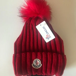 Röd ribbad mössa från Moncler - Snygg röd mössa från Moncler med en stor fluffig pälsboll på toppen. Mössan är ribbad och har Monclers logotyp framtill. Perfekt för att hålla värmen under kalla dagar.