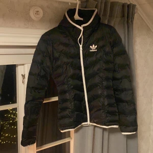 Svart dunjacka från Adidas - Snygg svart dunjacka från Adidas med vit kant och logga på bröstet. Jackan har en figurnära passform och långa ärmar. Perfekt för kyliga dagar. Den är i storlek XS/34, bra skick. Tror inte att just denna modellen finns att köpa längre. Den är runt 4 år gammal, men har bara legat på vinden senaste tiden. Säljes för 300+ frakt🌼