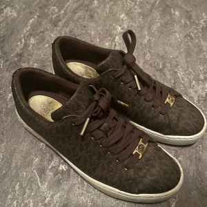Bruna sneakers från Michael Kors - Snygga bruna sneakers från Michael Kors med monogrammönster och gulddetaljer. Skorna har en vit sula och bruna snören med guldfärgade ändar. Perfekta för en stilren look.