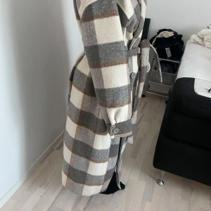 Rutig kappa med bälte - Snygg rutig kappa i beige, grå och brun med knytskärp i midjan. Långärmad design med knappar vid ärmsluten. Perfekt för höst och vår.