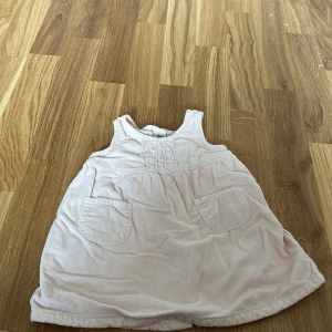 Ljusrosa ärmlös klänning för bebis - Söt ljusrosa ärmlös klänning för bebis med knappar på baksidan och två fickor framtill. Perfekt för varma dagar och enkel att ta på och av. Tillverkad i mjukt material för extra komfort.