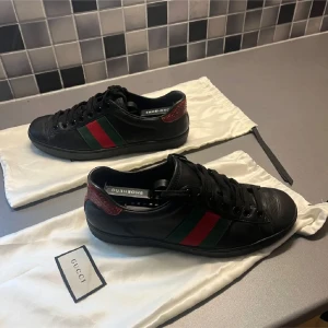 Svarta sneakers från Gucci - Storlek 41,5 passar 42eu Säljer ett par snygga svarta sneakers från Gucci i bra skick. De har de klassiska gröna och röda ränderna på sidan och en röd detalj vid hälen. Perfekta för både vardag och fest 