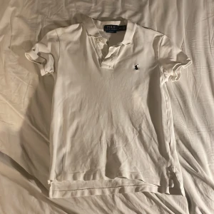 Vit pikétröja från Ralph Lauren - Säljer en klassisk vit pikétröja från Ralph Lauren i slim fit. Tröjan har korta ärmar och den ikoniska loggan broderad på bröstet. Perfekt för en stilren look.