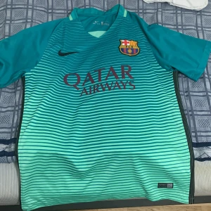 Turkos Barcelona fotbollströja från nike - Säljer en snygg turkos fotbollströja från Nike med FC Barcelona-logga och Qatar Airways-tryck. Tröjan är kortärmad och har ett randigt mönster i olika nyanser av turkos. Perfekt för fotbollsfans som vill ha något unikt! 🏆